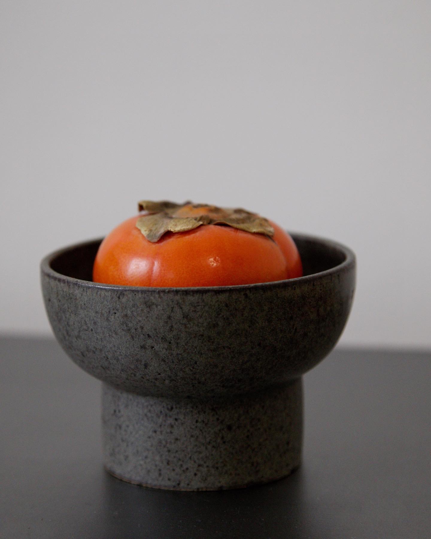 An orange persimmon resting in a dark ceramic bowl.